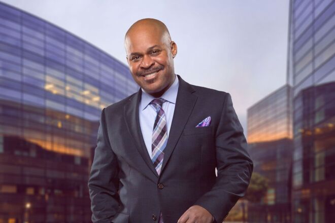 businessman in an urban portrat wearing grey suit and lavender shirt with office buildings in background. smiling with one hand in pocket