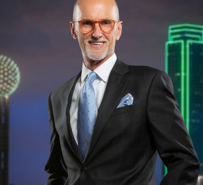 tulsa attorney in black suit with blue tie poses smiling with hands in pockets in front of Dallas skyline