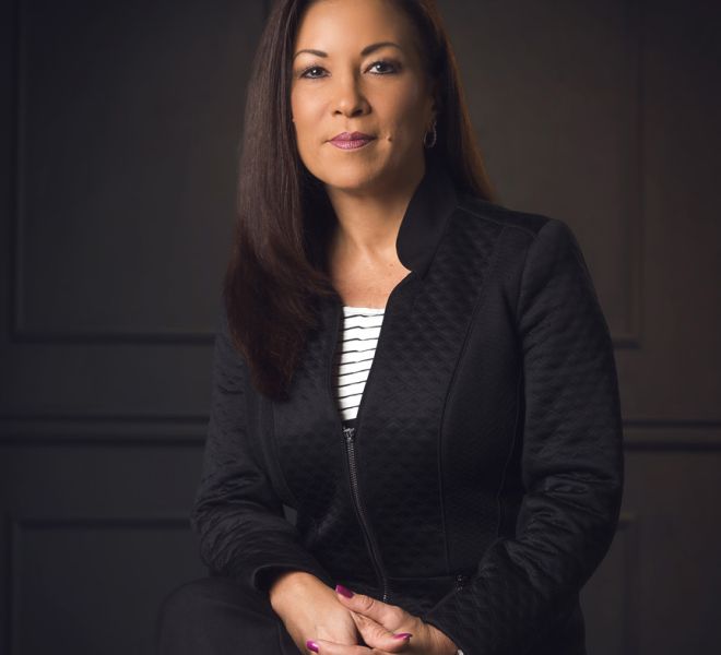 attractive businesswoman smiles and sits on a stool with hands clasped and legs crossed wearing a black jacket and black pants with panel background