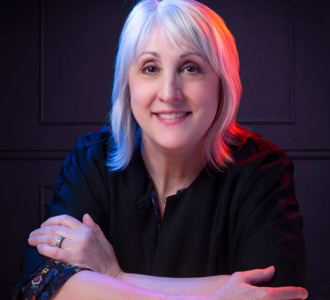 businesswoman backlit by red and blue lights with panel background poses with arms crossed on a backlit white tabletop wearing a black top with white hair