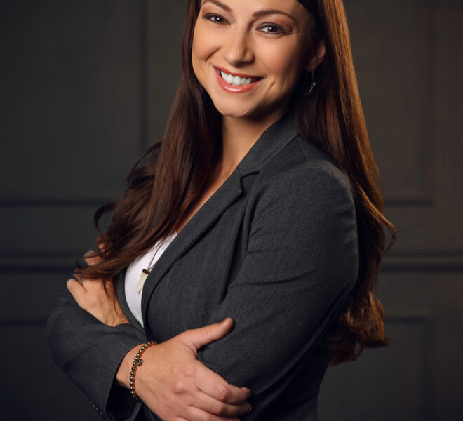 young professional woman with long auburn hair and grey suit smiles and crosses arms in front of wood panel background