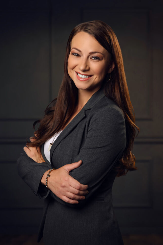 young professional woman with long auburn hair and grey suit smiles and crosses arms in front of wood panel background