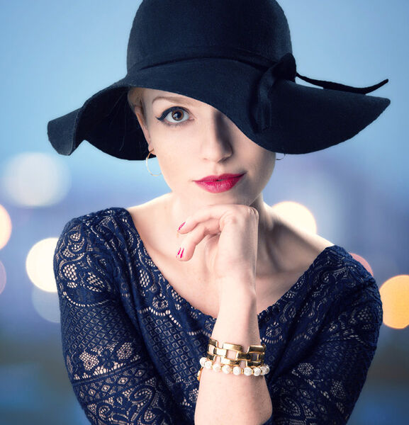 young woman with floppy stylish had covering one eye, wearing blue dress smiles sweetly with heavily blurred bokeh blue background.