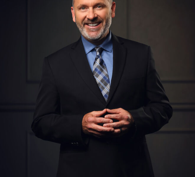 businessman in black suit and blue shirt poses standing in front of a wood panel background, smiling with hands together