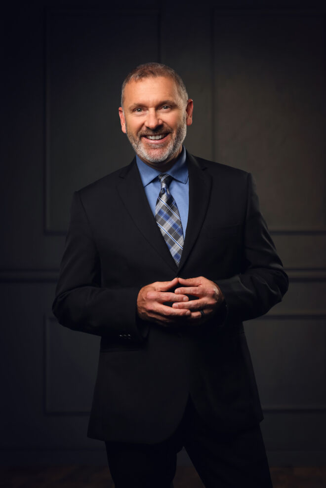 businessman in black suit and blue shirt poses standing in front of a wood panel background, smiling with hands together
