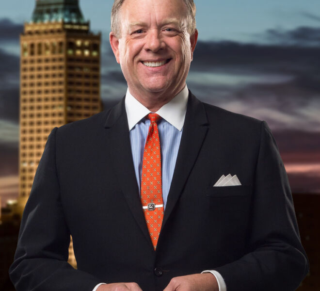 Attorney in lack suit and orange tie, poses with hands together, smiling in front of Tulsa skyline