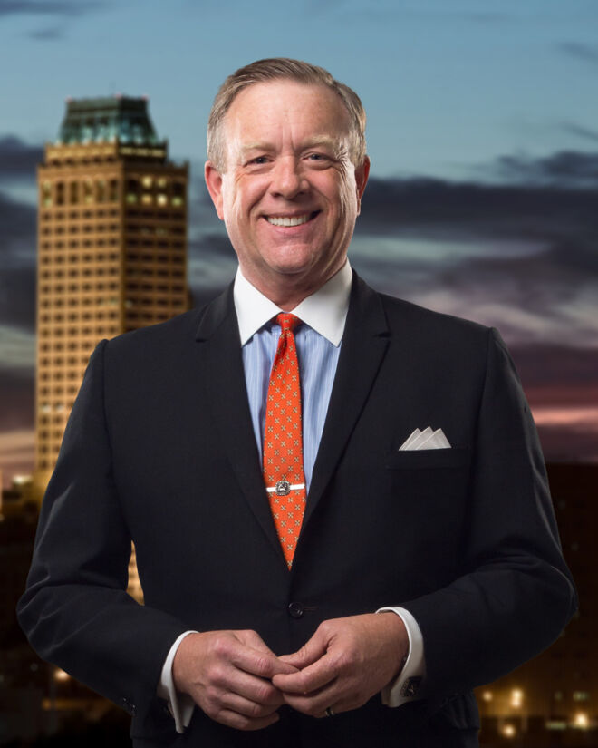 Attorney in lack suit and orange tie, poses with hands together, smiling in front of Tulsa skyline