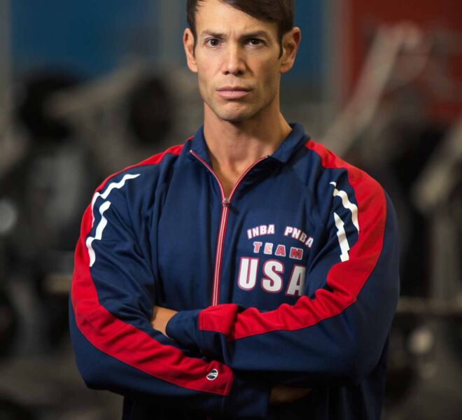 male fitness influencer in blue athletic pullover standing with serious look and arms crossed in gym with weight equipment behind him.