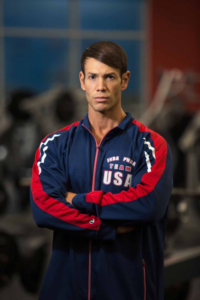male fitness influencer in blue athletic pullover standing with serious look and arms crossed in gym with weight equipment behind him.
