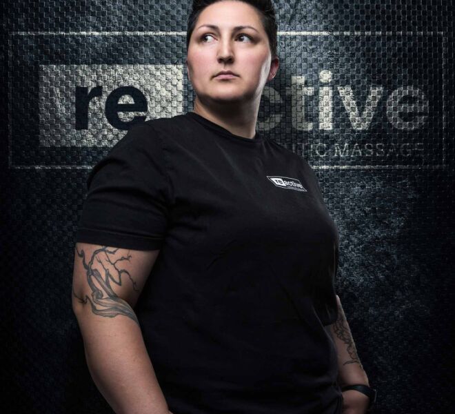 famale sports massage therapist with short hair and strong build stands in front of dark logo wall looking off camera.