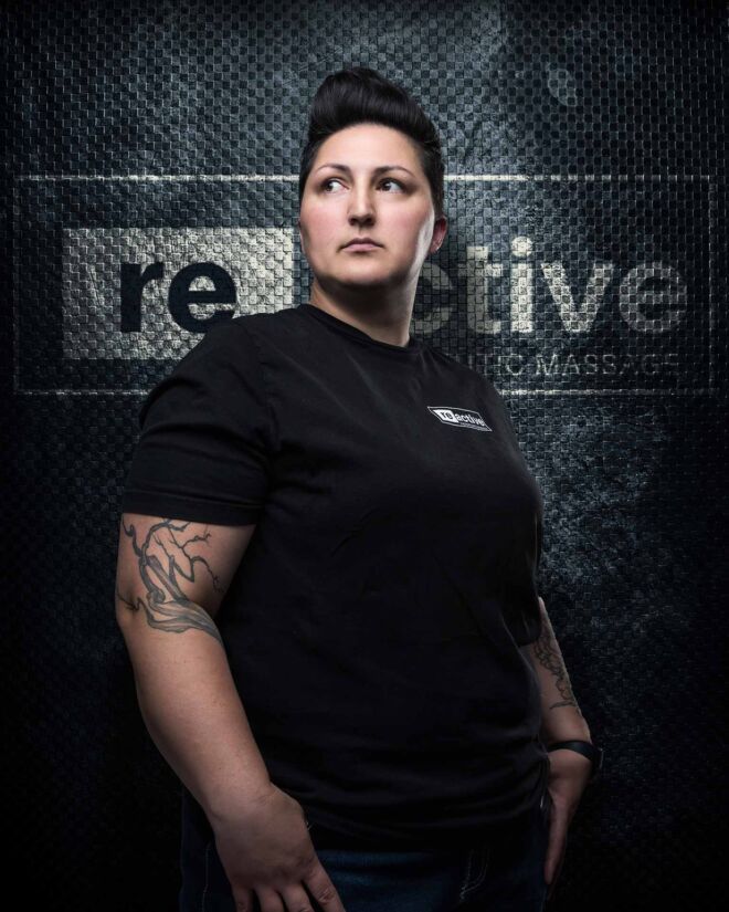 famale sports massage therapist with short hair and strong build stands in front of dark logo wall looking off camera.