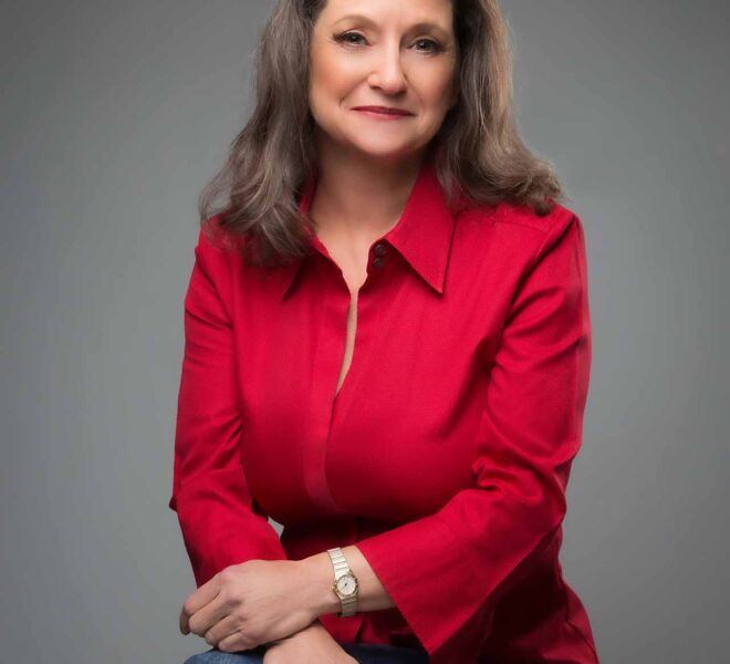 female doctor in casual jeans and red top sits on a stool with arms crossed over her knee, smiling on a grey portrait studio background