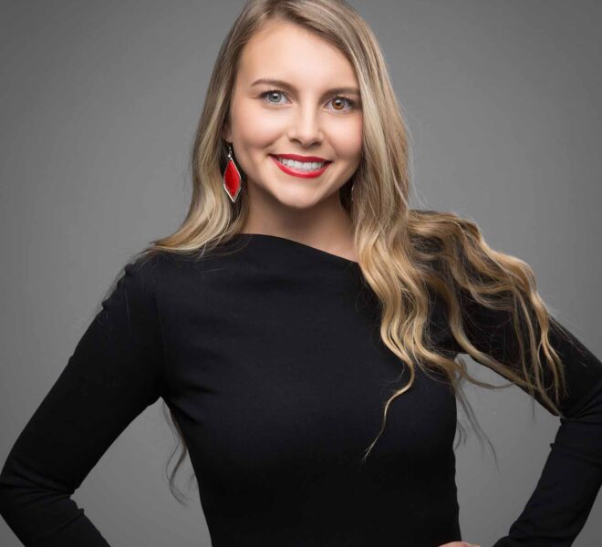 Pretty blonde realtor in black dress with different colored eyes smiles for headshot on grey background