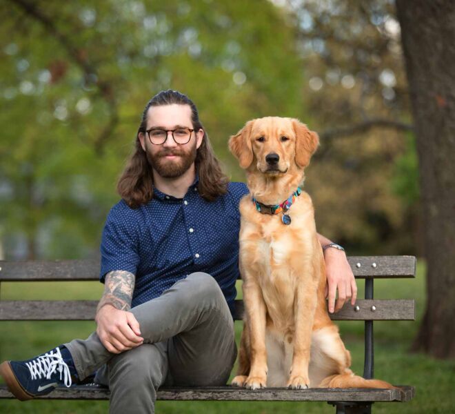young man in blue shirt with his arm around his golden retriever dog, as they both sit on a park bench posing for headshots.