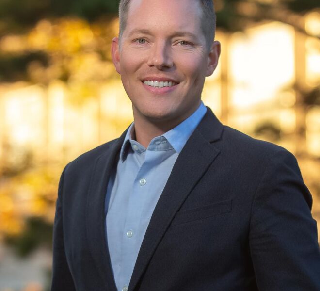 Tulsa professional man in a dark suit and light blue shirt poses smiling outdoors with blurred out fall foliage in the background