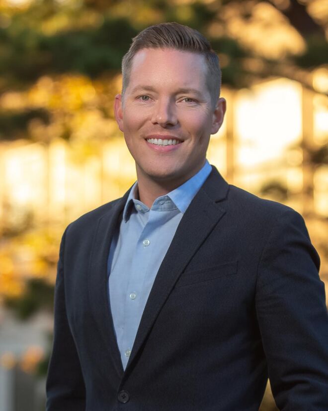 Tulsa professional man in a dark suit and light blue shirt poses smiling outdoors with blurred out fall foliage in the background