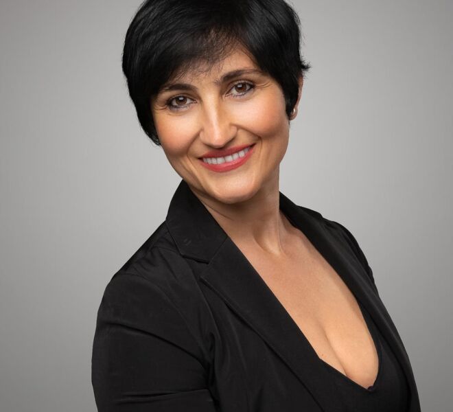 female realtor in a black dress and black jacket and short black hair poses smiling on a grey background.