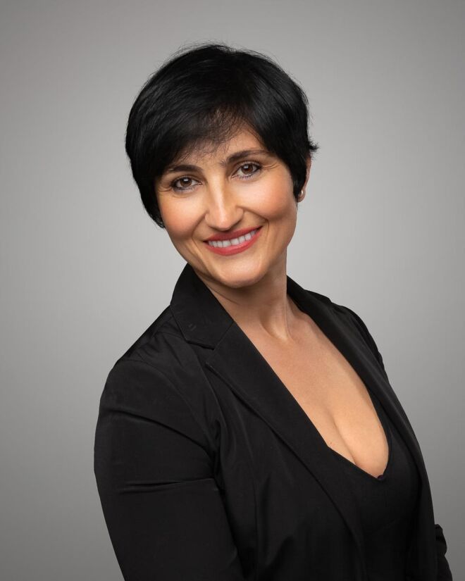 female realtor in a black dress and black jacket and short black hair poses smiling on a grey background.