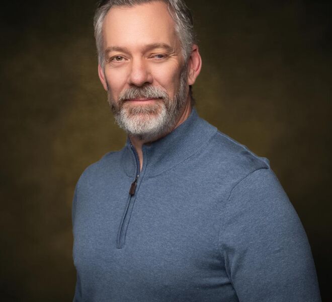 Tulsa attorney in a casual grey pullover poses in front of a brown portrait background smiling in a headshot