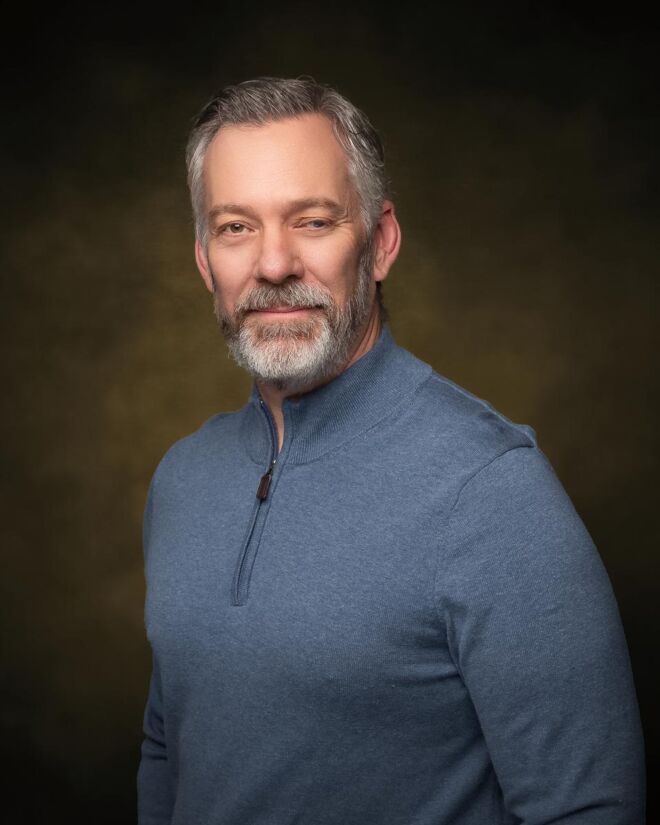 Tulsa attorney in a casual grey pullover poses in front of a brown portrait background smiling in a headshot