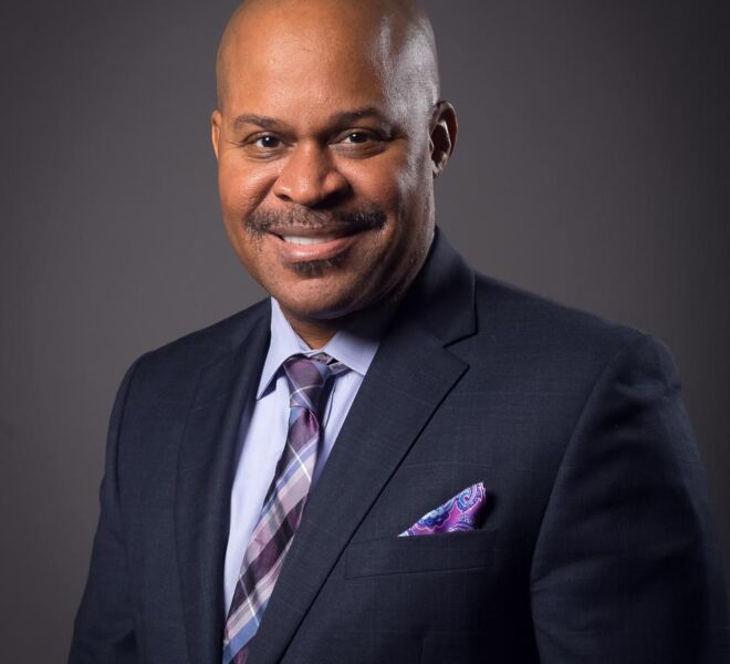african american businessman in a blue suit and lavender shirt poses for headshot on a grey background