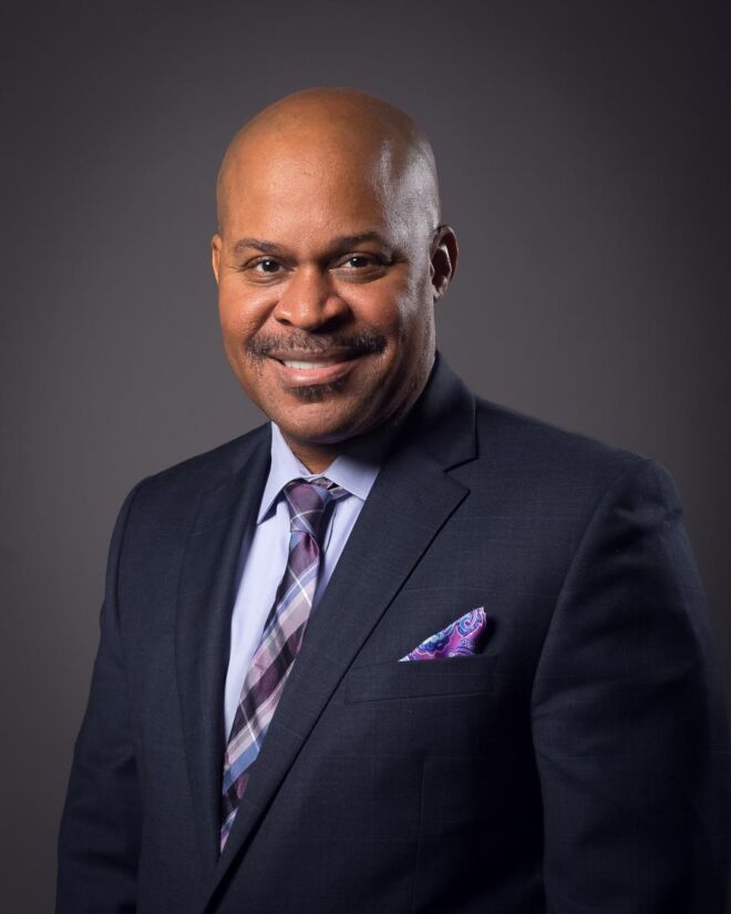 african american businessman in a blue suit and lavender shirt poses for headshot on a grey background