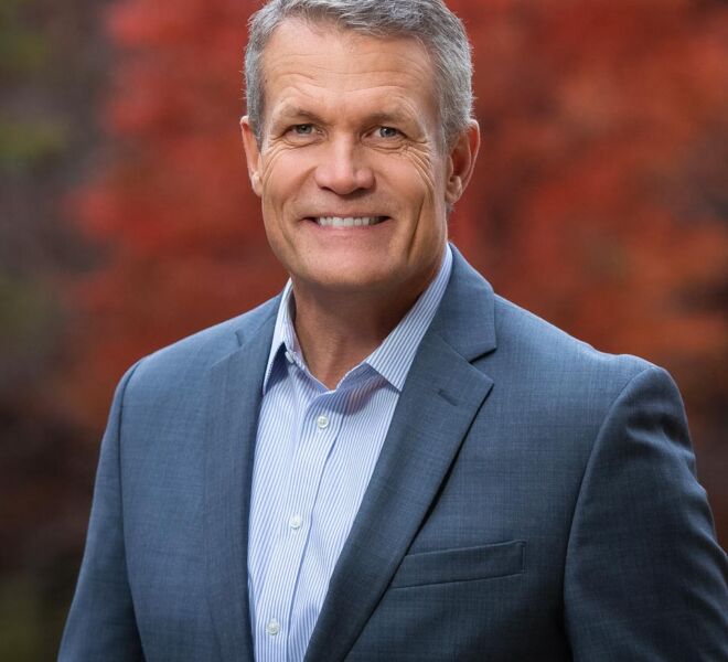 executive poses in tulsa park for headshots wearing a grey suit and blue and white striped shirt with fall colors in background