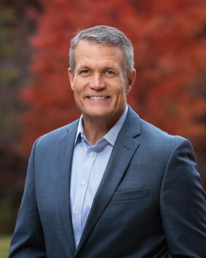 executive poses in tulsa park for headshots wearing a grey suit and blue and white striped shirt with fall colors in background