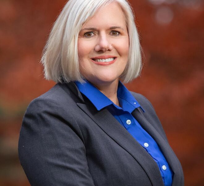 female executive in grey suit and blue top poses smiling outdoors in front of a Tulsa park fall foliage background