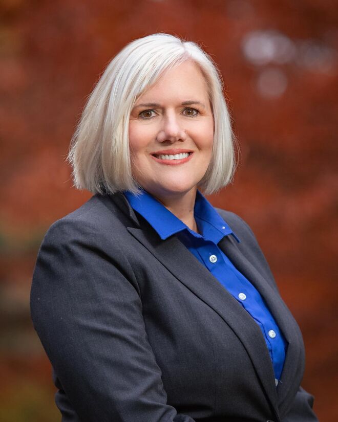 female executive in grey suit and blue top poses smiling outdoors in front of a Tulsa park fall foliage background
