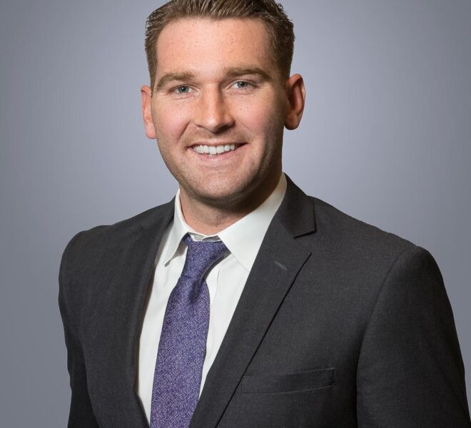 young male attorney with a dark grey suit and purple tie poses smiling in front of a grey background