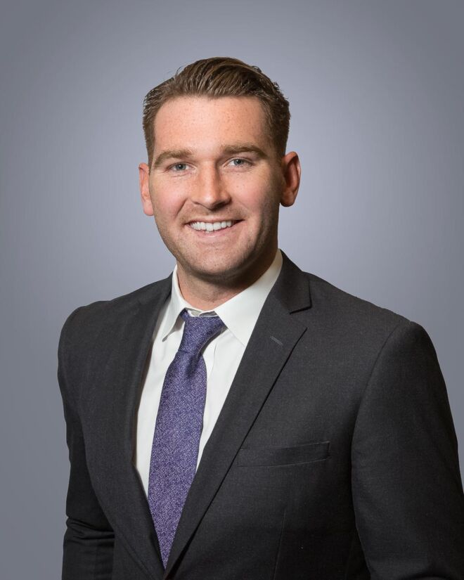 young male attorney with a dark grey suit and purple tie poses smiling in front of a grey background