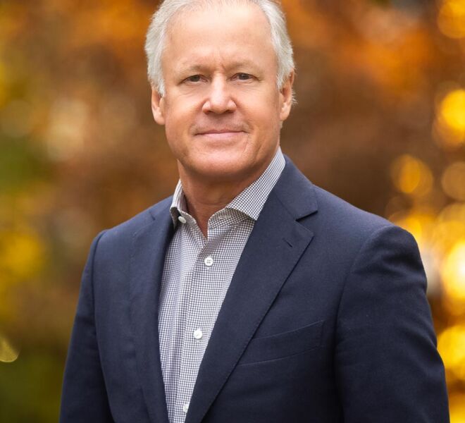 Executive businessman poses with a subtle smile wearing a blue jacket and white shirt with a fall foliage background