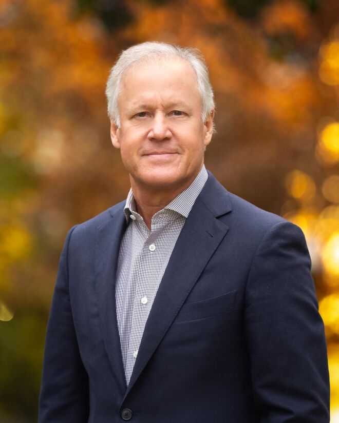 Executive businessman poses with a subtle smile wearing a blue jacket and white shirt with a fall foliage background