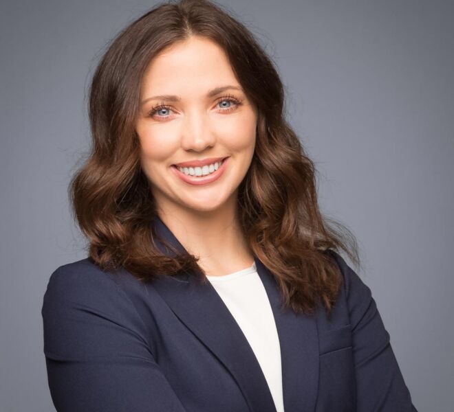young female attorney in a blue jacket and white top poses smiling with arms crossed on a grey background