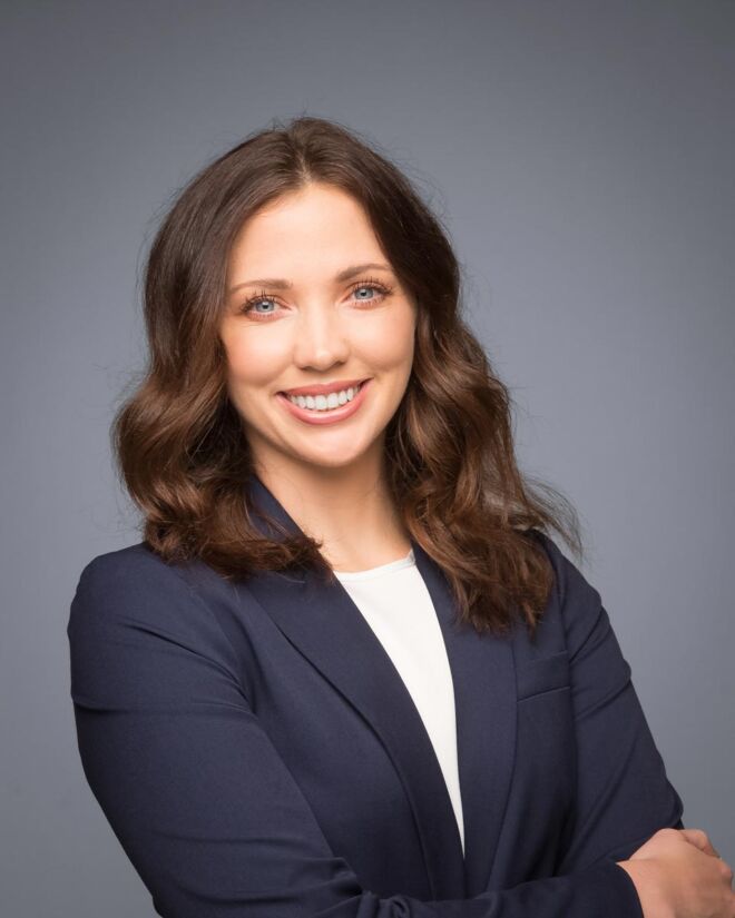 young female attorney in a blue jacket and white top poses smiling with arms crossed on a grey background