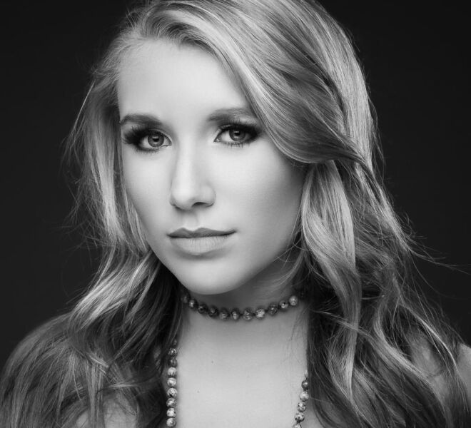 black and white headshot of a beautiful young female singer with long blond hair, wearing a serious look and a pearl necklace on a dark background