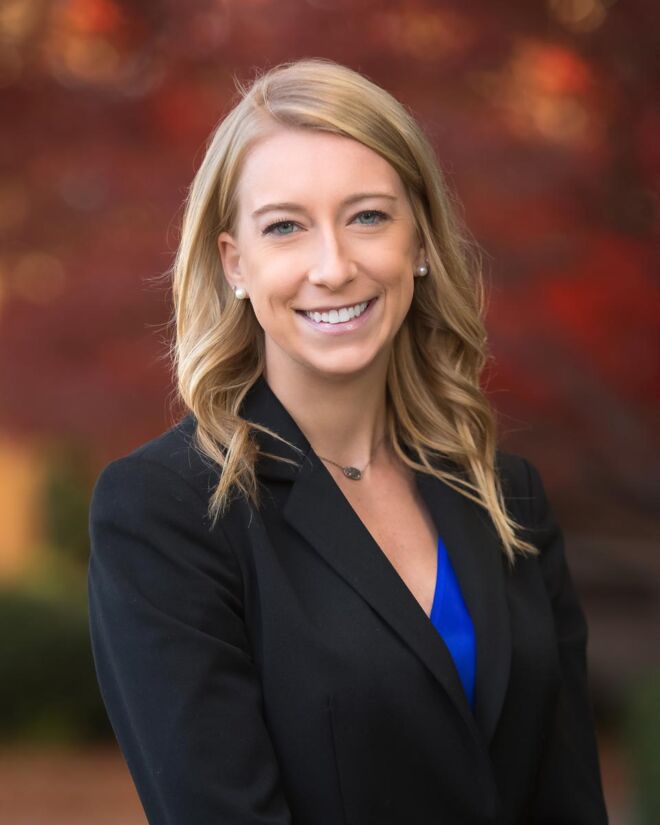 young blonde female attorney wearing a black suit with a blue top, poses outdoors smiling for a photo with fall foliage in the background