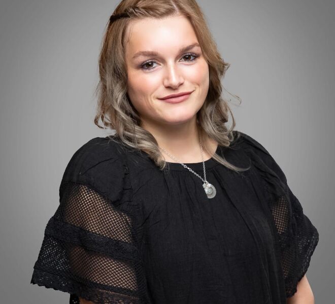 young professional woman with blonde hair and a black dress smiles for a photo on a grey background