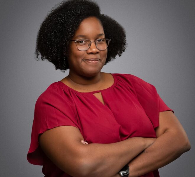 Young african american woman in glasses smiles with her arms crossed, wearing a red top on a grey background