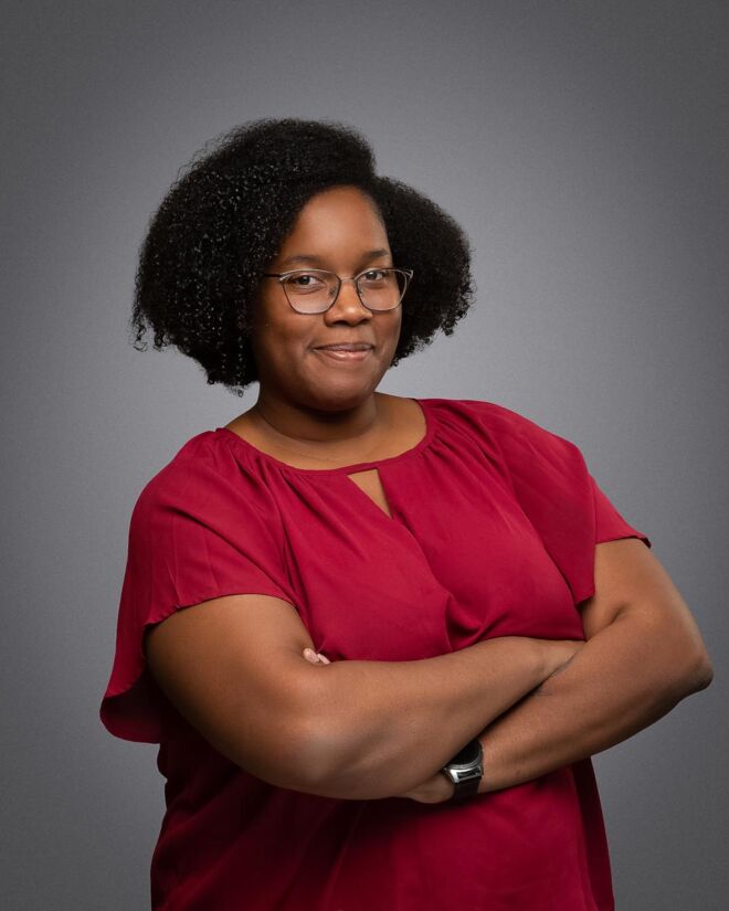 Young african american woman in glasses smiles with her arms crossed, wearing a red top on a grey background