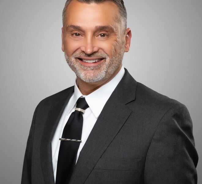 male tulsa realtor with dark hair and a white beard, wearing a black tie and grey suit poses for his headshot on a grey background