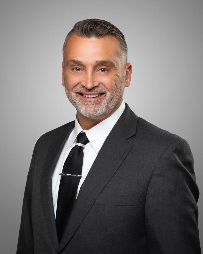 male tulsa realtor with dark hair and a white beard, wearing a black tie and grey suit poses for his headshot on a grey background