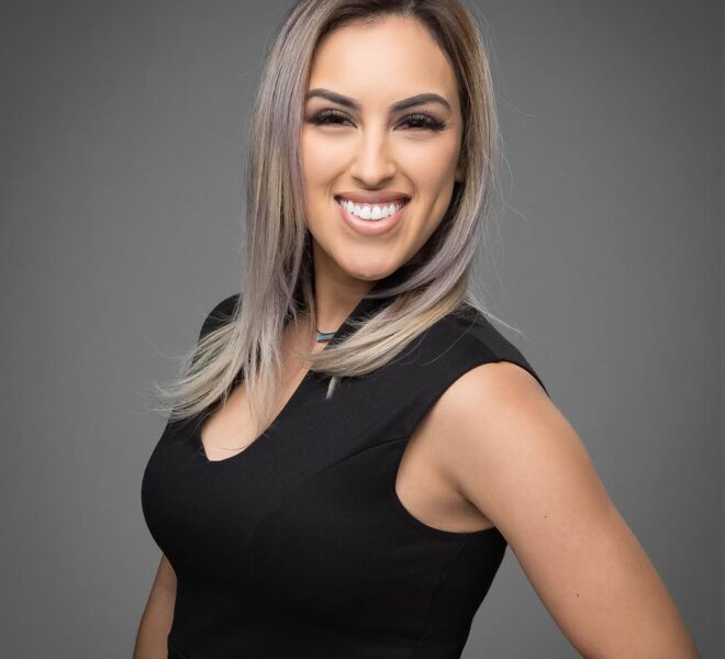 pretty young female Tulsa realtor poses with her hand on her hip, in a black dress with blond hair and pink highlights, smiling for a headshot on a grey background