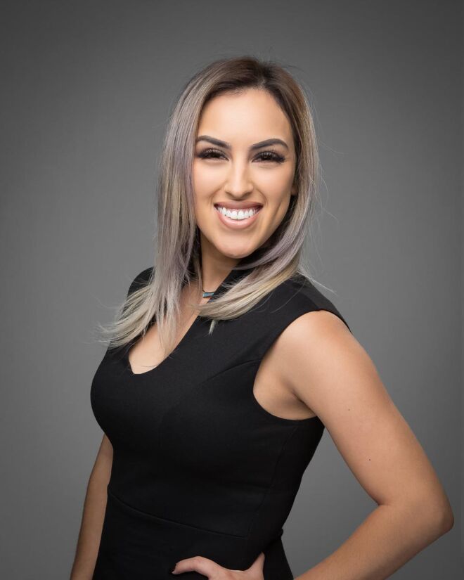 pretty young female Tulsa realtor poses with her hand on her hip, in a black dress with blond hair and pink highlights, smiling for a headshot on a grey background