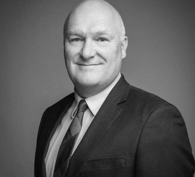 black and white headshot portrait with an older balding businessman in a dark suit on a medium tone background
