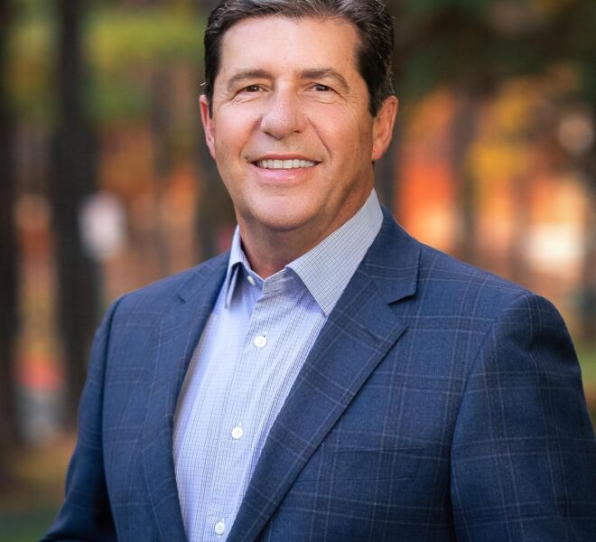 Businessman in a blue blazer and light blue shirt poses smiling outdoors in a Tulsa park setting with fall colors