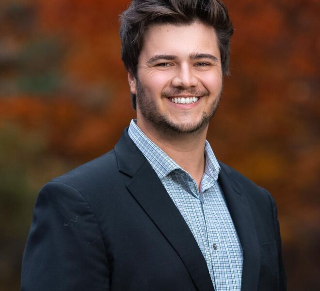 handsome young businessman in a blue jacket and checkered white shirt poses smiling for headshots in front of fall foliage outdoors