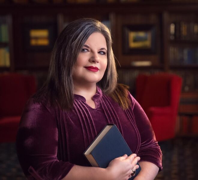 Author poses in Tulsa mansion library for her headshot, holding a book and wearing a plumb colored dress