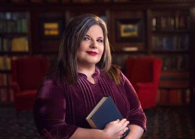 Author poses in Tulsa mansion library for her headshot, holding a book and wearing a plumb colored dress